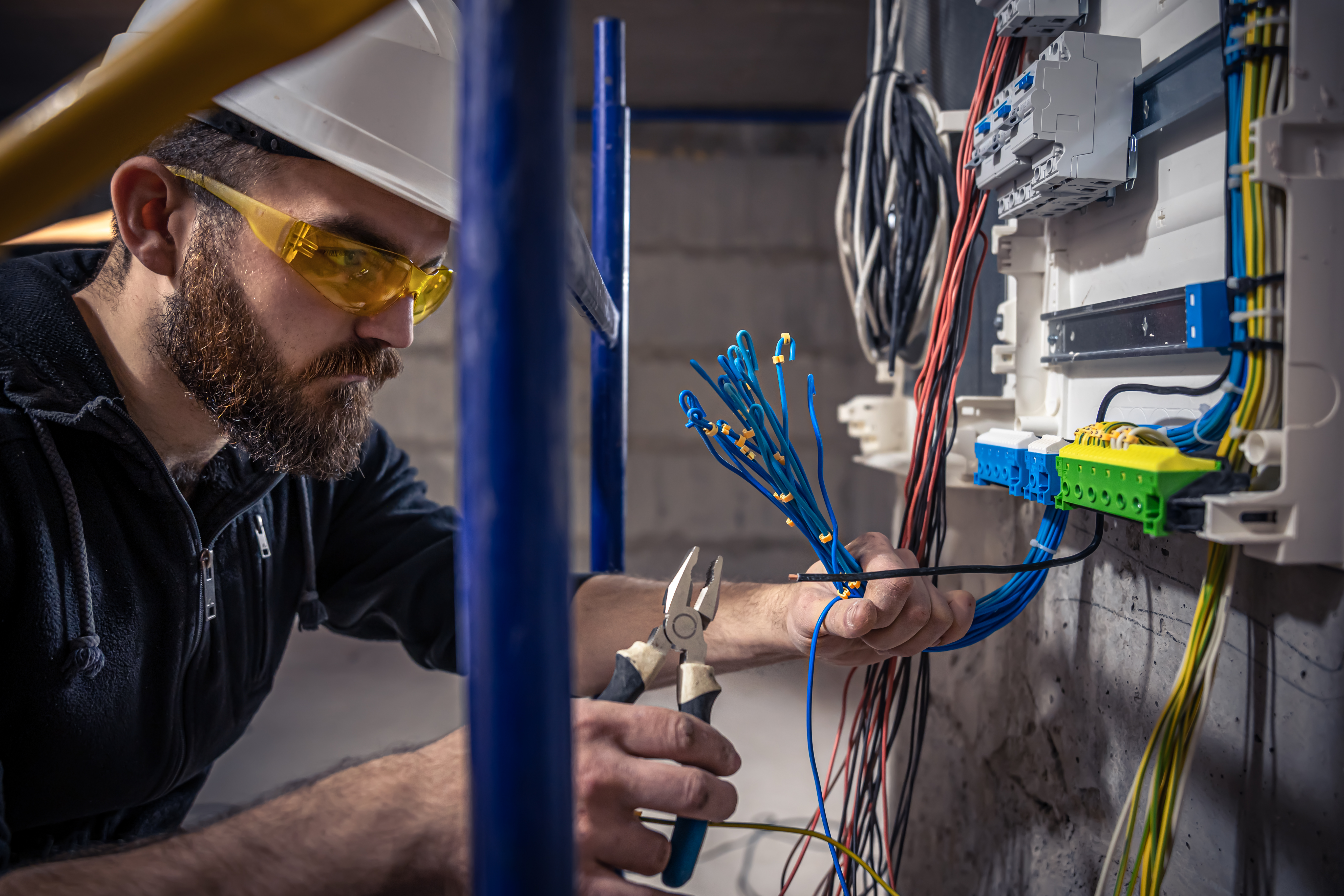 A male electrician works in a switchboard with an electrical connecting cable, connects the equipment with tools.