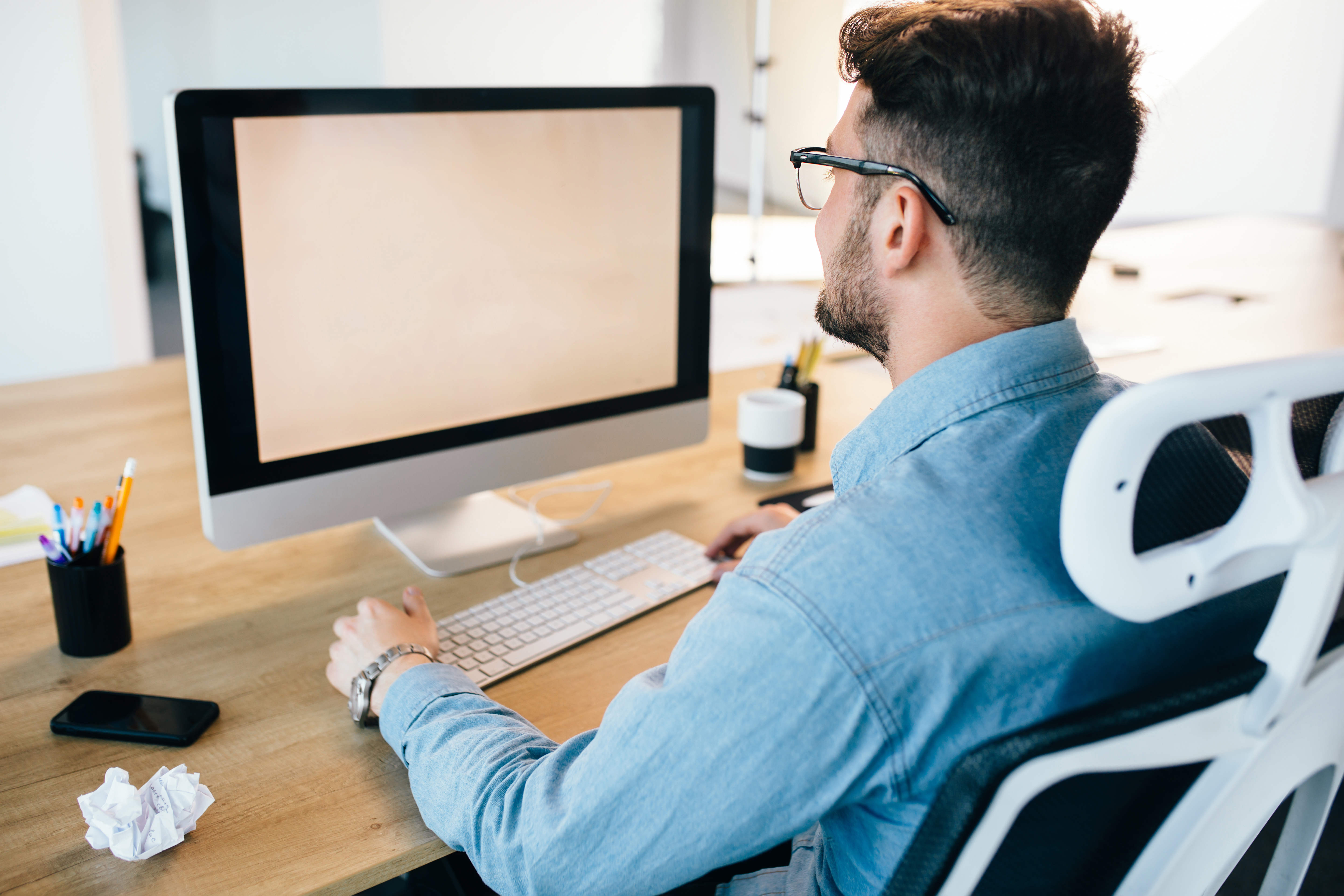 Young dark-haired man is working with a computer at his desktop in office. He wears blue shirt and looks busy. View from back.
